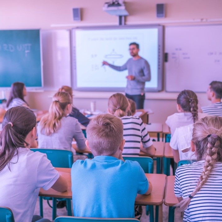 fotografia de um professor ministrando aula em uma sala equipada com lousa digital, enquanto alunos assistem atentamente.